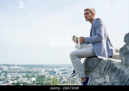 uomo d'affari con un tablet in mano siede su una roccia sullo sfondo della città e guarda in lontananza Foto Stock