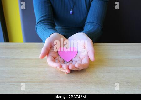 Il cuore di carta rosa si trova nelle palme aperte di una ragazza in un maglione blu. Foto Stock