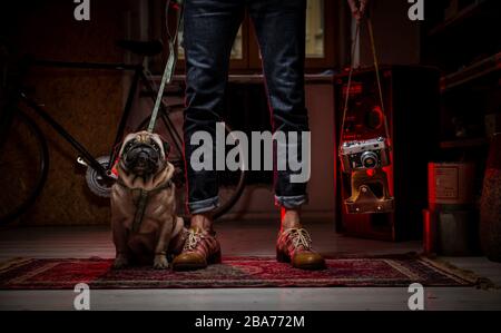 Un uomo hipster che stnding sul tappeto nella stanza che tiene un cane pug su un guinzaglio in una mano e vecchia macchina fotografica vintage nell'altra mano. Bicicletta sul retro Foto Stock