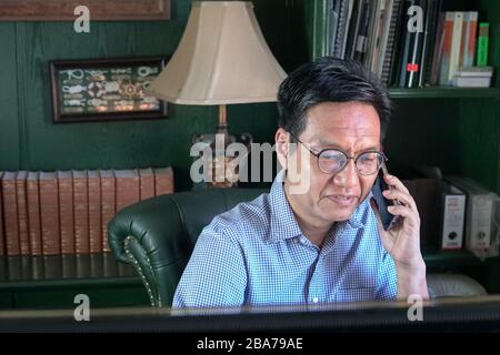Concetto di lavoro da casa. Uomo d'affari cinese asiatico sul telefono cellulare nella sua stanza di studio. Foto Stock