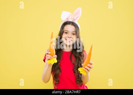 Tradizione primaverile. Orecchie conigliate per bambini. Dieta per la salute. Beneficio di mangiare la carota. Carota di Pasqua. Ragazza tenere carota. Gioca con il cibo. Bambino sfondo giallo. Le vacanze di Pasqua stanno arrivando. Cibo sano. Foto Stock