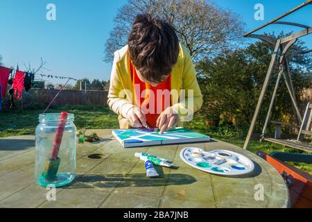 Un ragazzo che fa un dipinto su un tavolo nel giardino posteriore in una giornata di sole. Foto Stock