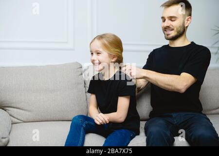 Giovane padre a casa con la sua bambina carina che la rende un hairstyle. Outfit casual simile - t-shirt e jeans neri. Foto Stock