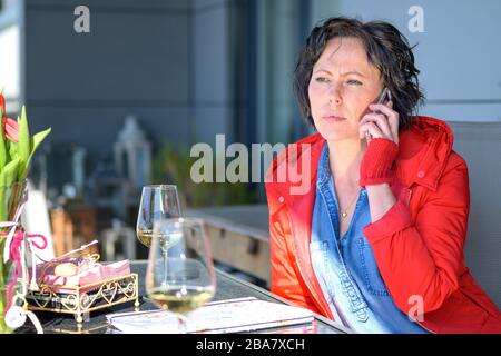 Donna alla moda che chiacchiera sul suo cellulare a un tavolo del ristorante mentre attende il suo ordine di cibo guardando da parte con un look attento Foto Stock