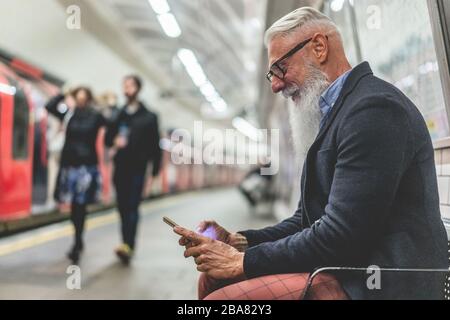 Uomo di hipster anziano che usa lo smartphone nella metropolitana - persona matura di moda che si diverte con le tendenze tecnologiche che aspettano il suo treno - anziani gioiosi Foto Stock