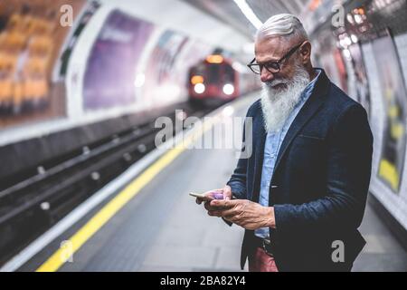 Uomo di hipster anziano che usa lo smartphone nella metropolitana - persona matura di moda che si diverte con le tendenze tecnologiche che aspettano il suo treno - anziani gioiosi Foto Stock