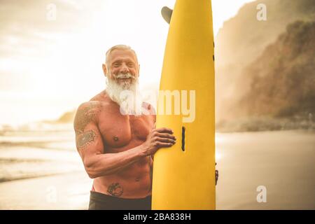 Tattooed surfer senior tenendo surf board sulla spiaggia al tramonto - ragazzo felice vecchio divertirsi facendo sport estremo - gioioso anziano concetto - Focus on hi Foto Stock