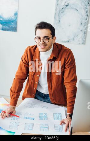 Bel progettista ux sorridente alla macchina fotografica mentre lavora con i modelli di Web site al tavolo Foto Stock