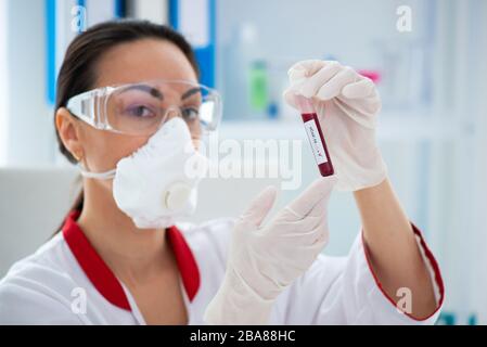 Dottore donna in maschera con un pallone con un campione di sangue. Ricerca di laboratorio e rilevamento del coronavirus Covid-19 Foto Stock