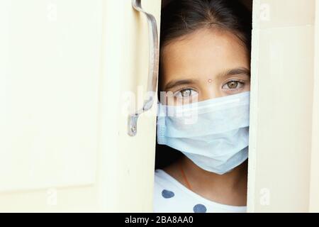 Giovane ragazza triste con maschera medica che indossa sneaking fuori attraverso la porta di casa - concetto di quarantena casa a causa di covid-19 o scoppio di coronavirus. Foto Stock