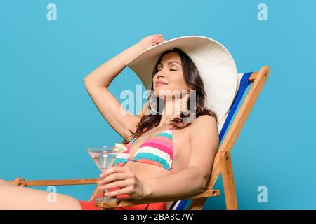 donna attraente e felice che prende il sole sulla sedia a sdraio mentre tiene un bicchiere di cocktail su sfondo blu Foto Stock