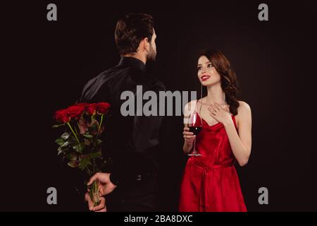 Elegante uomo nascosto bouquet di rose vicino ragazza sorridente con vino rosso isolato su nero Foto Stock