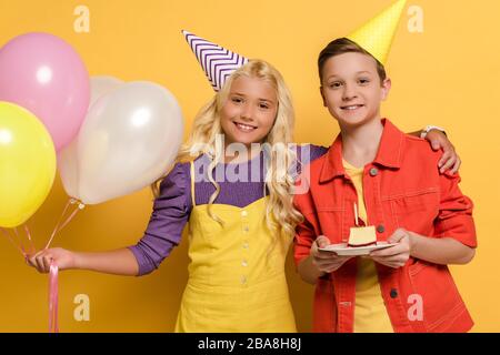 bambini sorridenti con cappellini da festa che tengono palloncini e piatto con torta di compleanno su sfondo giallo Foto Stock