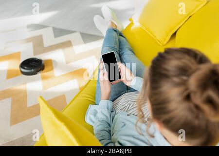 vista dall'alto di una donna che tiene uno smartphone con schermo vuoto mentre l'aspirapolvere robot lava il tappeto nel soggiorno Foto Stock