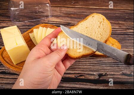 Qualcuno spalma il burro con un coltello su pane tostato di grano fresco, burro e formaggio in un tegame di legno su uno sfondo di legno. Primo piano Foto Stock