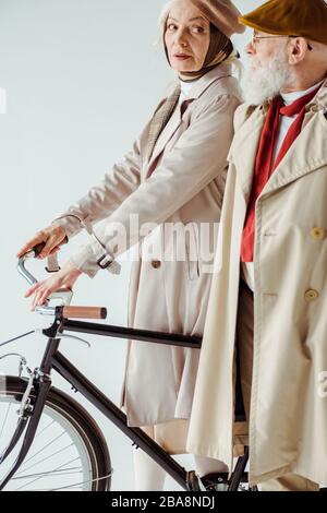 Vista laterale di elegante donna anziana con bicicletta guardando uomo elegante in trench isolato su bianco Foto Stock