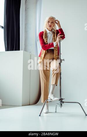 Elegante donna anziana in piedi vicino treppiede su sfondo bianco in studio fotografico Foto Stock