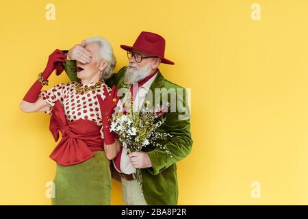 Uomo anziano elegante che copre gli occhi alla donna e tenendo bouquet di fiori selvatici su sfondo giallo Foto Stock