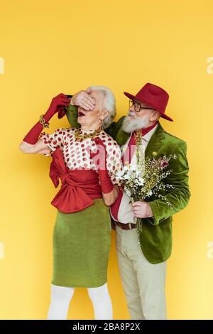 Bell'uomo anziano che copre gli occhi alla donna alla moda e tenendo bouquet di fiori selvatici su sfondo giallo Foto Stock