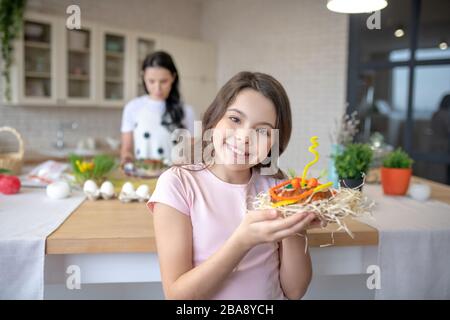 Ragazza sorridente in jeans in piedi in cucina e tenendo la pasqua mordente nelle sue mani Foto Stock