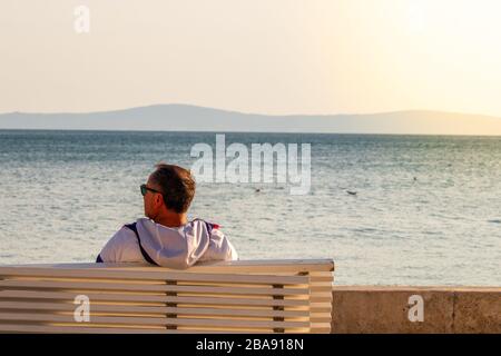 Uomo più anziano visto da dietro seduto su una panchina bianca sulla riviera di Spalato, Croazia. Godendo della vista del mare, gabbiani che volano sul mare Foto Stock