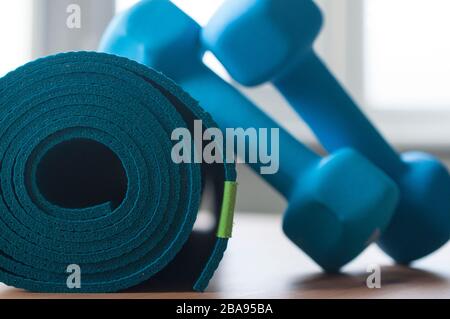 Un tappetino di yoga arrotolato blu. Due manubri blu da 2 kg appoggiati su di esso. Attrezzature per il fitness per l'allenamento domestico e la flessibilità. Foto Stock