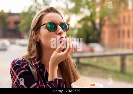 Una giovane ragazza mangia le sue patatine fritte proprio sulla strada. È molto affamata, appetito brutale svegliato in lei. Foto Stock