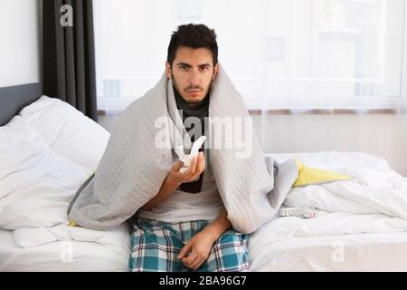 Un giovane è seduto sul suo letto. È malato, ha una temperatura corporea elevata, naso che cola e tosse. Foto Stock
