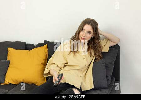 Una donna siede a casa sul divano e guarda la TV. Sta tenendo un telecomando, cercando di trovare un canale interessante. Foto Stock
