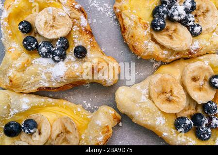 Biscotti a base di pasta sfoglia con budino, banana e mirtilli cosparsi di zucchero in polvere adagiato sulla carta da forno. Foto Stock