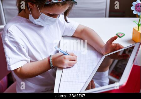 Formazione online a distanza. Studentessa di malattia in maschera medica che studia a casa con un tablet digitale in mano e che fa i compiti scolastici. Libri di formazione e notebook a tavola. Messa a fuoco morbida Foto Stock