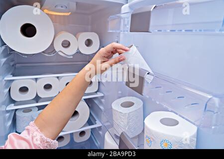 La mano femminile prende un rotolo di carta igienica dalla porta del frigorifero. Panico che acquista carta igienica in tutti i paesi durante la diffusione del coronavirus COVID-19 e la quarantena. Concetto Covidiot Foto Stock