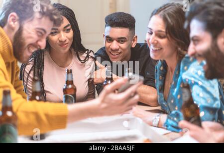 Gruppo di giovani sorridenti che guardano video su smartphone e bevono birre in una casa. Amici multirazziali che si divertono con lo smartphone, social netw Foto Stock