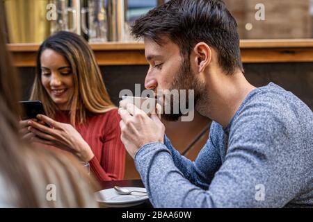 Gruppo di amici al bar. Giovane adulto bearded che beve cappuccino da una tazza bianca. Altri amici sorridenti guardando i loro smartphone. Outdo Foto Stock