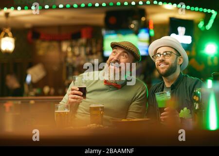 Uomini sorridi bearded bere birra e guardare sport mentre si siede nello sport bar Foto Stock