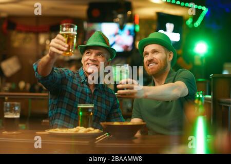 Due amici in cappelli verdi che alzano i loro bicchieri di birra mentre guardano la partita di sport nel pub Foto Stock