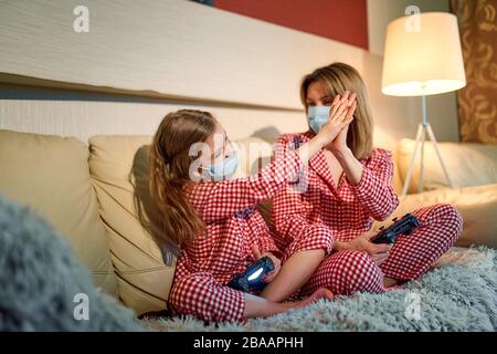 Donna e giovane ragazza che indossano pigiama e maschera medica protettiva seduta sul divano in soggiorno con controller di videogiochi a casa isolamento auto quarantena, covid-19. Foto Stock