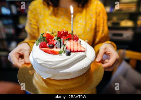 Immagine a profondità di campo bassa (fuoco selettivo) con una donna che tiene una torta di meringa di frutta fresca con una candela accesa. Foto Stock