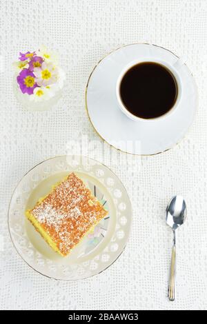 Torta di ravani greco con una tazza di caffè nero, un cucchiaio e un piccolo vaso con fiori primula sullo sfondo di pizzo Foto Stock