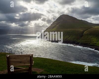 Isole Faroe. Villaggio di Gjógv. Vista dalla collina sopra il villaggio. Panca in legno in primo piano. La baia sullo sfondo. Cielo drammatico con sole b Foto Stock