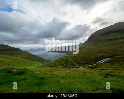 Isole Faroe. Vista sulla strada che porta a Funnings komuna. Campi verdi in primo piano e la baia sullo sfondo. Paesaggio frizzante. Foto Stock