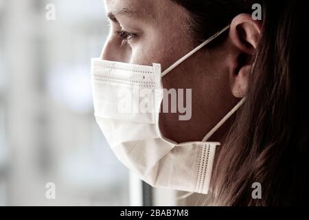 Particolare della giovane donna caucasica che indossa una maschera medica bianca per il viso per prevenire l'influenza. Mettere a fuoco sulla parte anteriore del viso, sfondo sfocato. Coronavirus, quarantena COVID-19. Concetto medico, infermiere. Foto Stock