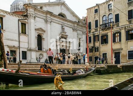 Una gondola legata nel canale con un gondoliere che aspetta nelle vicinanze. Un gruppo turistico si riunisce presso la chiesa. NMR Foto Stock