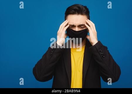 Ritratto di giovane malato in maschera nera protettiva sul viso che soffre di mal di testa e debolezza, isolato su sfondo blu. Sintomi tipici di Foto Stock