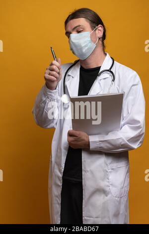 Medico che guarda ansioso di una penna che si chiede se ha toccato il suo viso con esso e potrebbe essere contaminato con sars-cov-2. Vestendo il accappatoio, lo stetoscopio e il proto Foto Stock