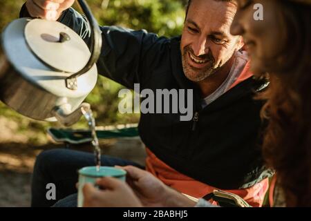 Coppia con caffè mentre campeggio in natura. Uomo che versa il caffè nella tazza della donna. Foto Stock