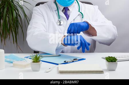 il medico in un cappotto medico bianco siede ad un tavolo in una sedia di pelle marrone e mette sui guanti medici sterili nelle sue mani prima di ricevere ed esaminare Foto Stock