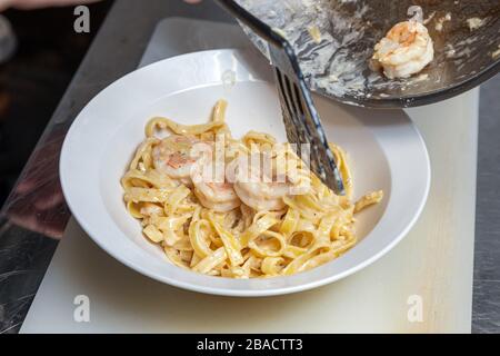 Lo chef Сlose-up maschile mescola grandi gamberi e pasta con salsa di crema da una padella a un piatto. Il processo di preparazione della pasta ai gamberi Foto Stock