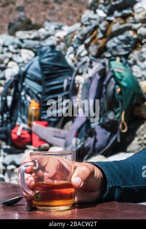 Primo piano di una tazza con tè in mano dell'escursionista. L'uomo di trekking beve un tè caldo nell'Himalaya Nepal. Concetto di trekking con zaini sullo sfondo. Foto Stock