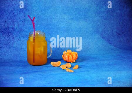 Una tazza di succo di mandarino con ghiaccio e cannucce accanto a pelati e tutta la frutta e fette di mandarino su uno sfondo blu. Close-up. Foto Stock